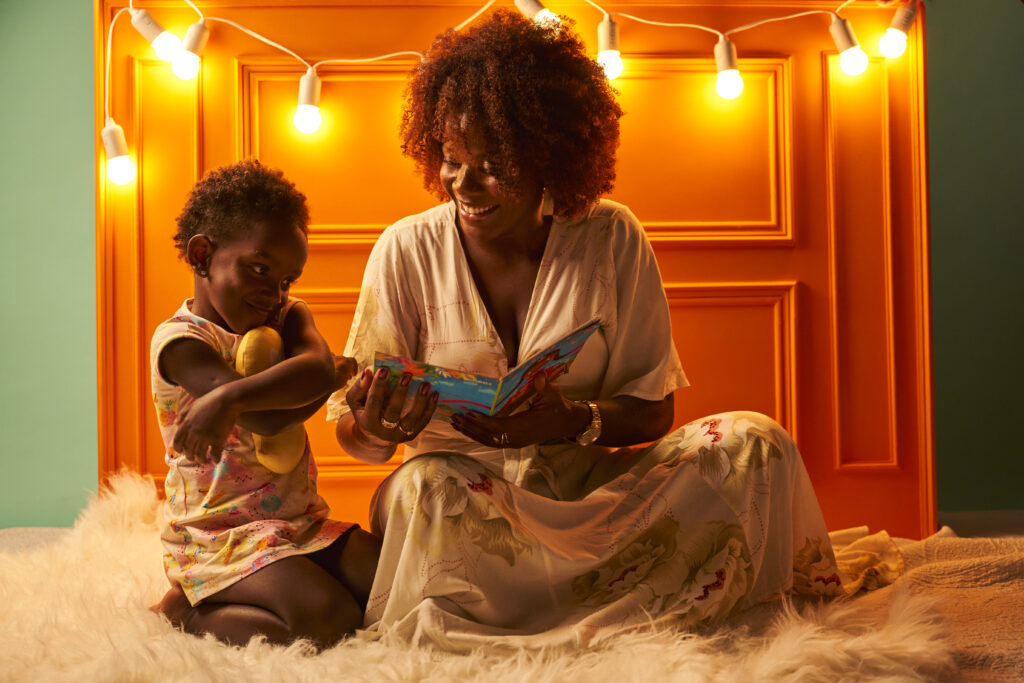 A imagem mostra uma mulher sorrindo enquanto lê para uma criança. 