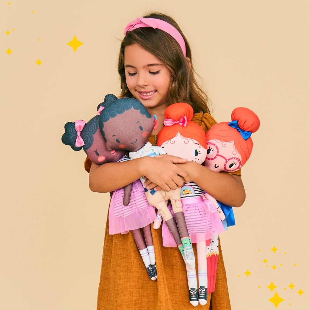 A imagem mostra uma menina vestida com um vestido infantil em tom terroso e segurando várias bonecas de pano da Alphabeto. 